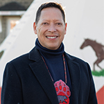 John Sylliboy, smiling in front of a white tipi decorated with a red pattern and an image of a horse.