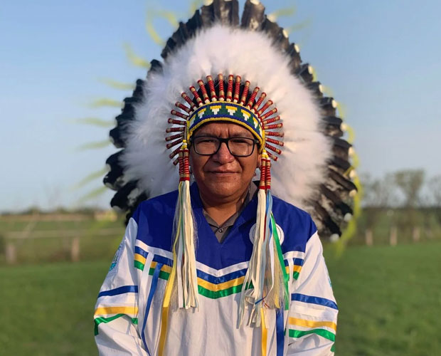 Grand Chief Scott Harper standing in an open field wearing a white shirt with white, blue, yellow and green ribbons on it. He is also wearing glasses and a feather headdress. 