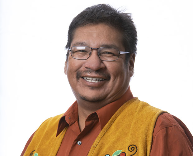 Le chef Wayne Moonias devant un fond blanc, vêtu d'une chemise orange et d'une veste à franges perlée de fleurs.