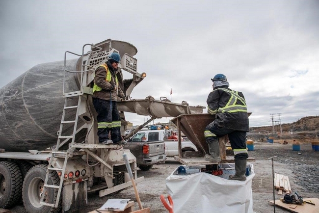 A construction worker stands on a platform on a cement truck directing the blow of backfill material. Another construction worker stands on a platform and holds a large metal funnel delivering the material into the underground. Work trucks are visible in the background.