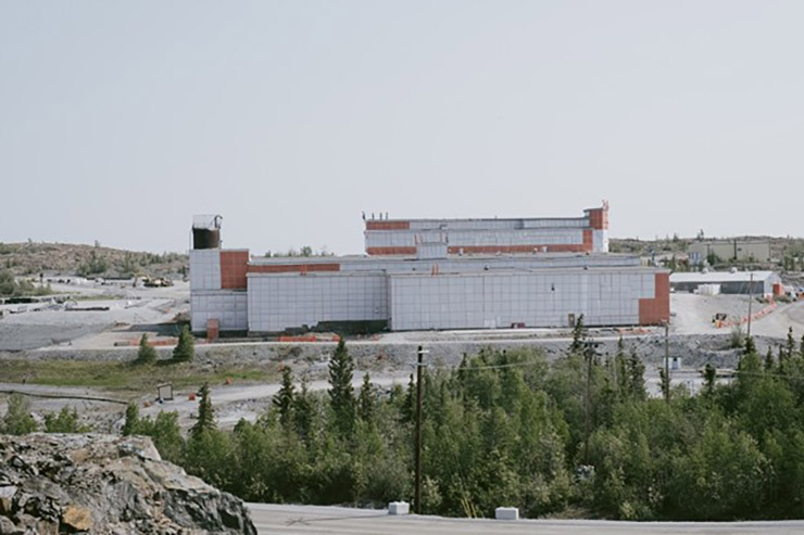A large industrial building wrapped in plastic on a construction site with rocks and trees in the foreground.