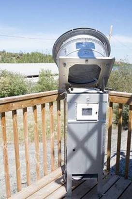 Photograph of air quality monitoring equipment with the top is opened to read instrumentation. The equipment is on a wooden deck with trees in the background.
