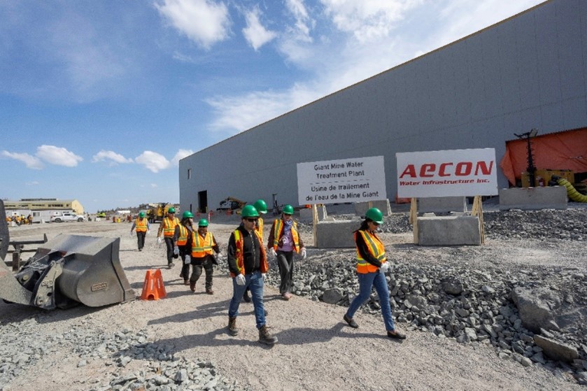Un groupe de personnes marche sur un chemin de gravier à côté d'une pelleteuse à l'extérieur d'un grand bâtiment industriel.