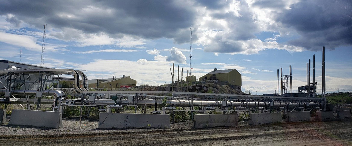 Une zone industrielle du site de la mine Giant montrant des tuyaux verticaux et des thermosiphons, qui sont de longs tuyaux verticaux remplis de dioxyde de carbone. Les bâtiments sont visibles sur un affleurement rocheux en arrière-plan de la photo.
