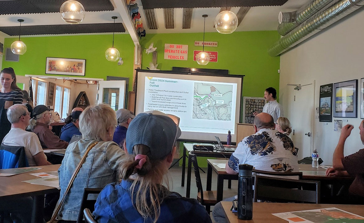A member of the Giant Mine Remediation Project team is shown presenting on a screen in front of a group of community members sitting at tables. The projected slide shows writing next to a map.