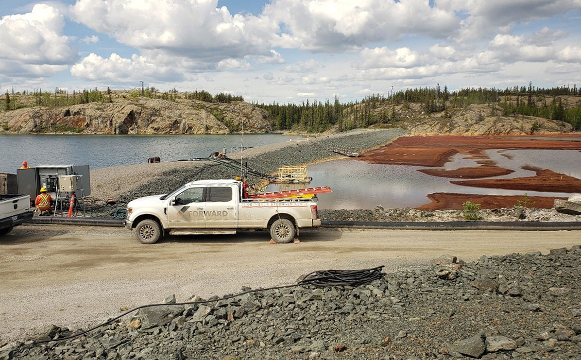 Une camionnette blanche est stationnée devant 2 bassins séparés par une berme de gravier. Le bassin de polissage se trouve à gauche et contient de l'eau claire, tandis que l'étang de décantation se trouve à droite et est partiellement recouvert de sulfate ferrique, le produit utilisé pour traiter l'eau, qui est de couleur rougeâtre.