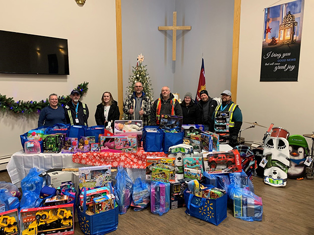 A group of 8 adults stand behind a table overflowing with unwrapped toys for a Christmas toy drive.