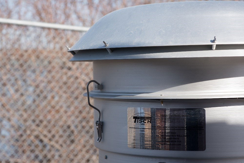 A metal cylinder that holds air quality monitoring equipment, with a chain link fence visible behind it.