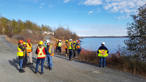 Un groupe de personnes vêtues d'un équipement de protection individuelle (EPI) se tient devant un lac et de vieilles maisons délabrées dont les fenêtres sont barricadées.