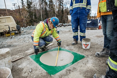 L'image montre une femme portant un EPI, qui mesure un échantillon de béton à la mine Giant, tandis que trois travailleurs portant un EPI observent l'essai.